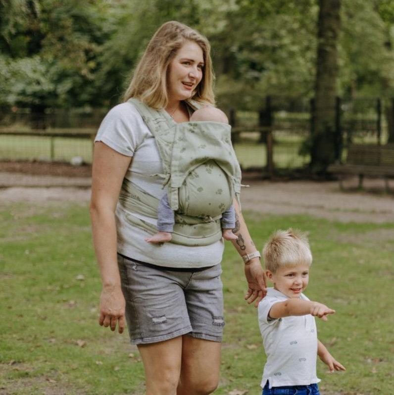 Mulher usando o Mei Tai Fidella FlyClick Toddler Clover verde claro, caminhando ao lado de uma criança pequena.