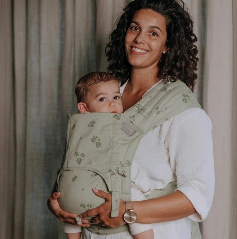 Mulher sorridente segurando um bebé num porta-bebés verde com padrão.