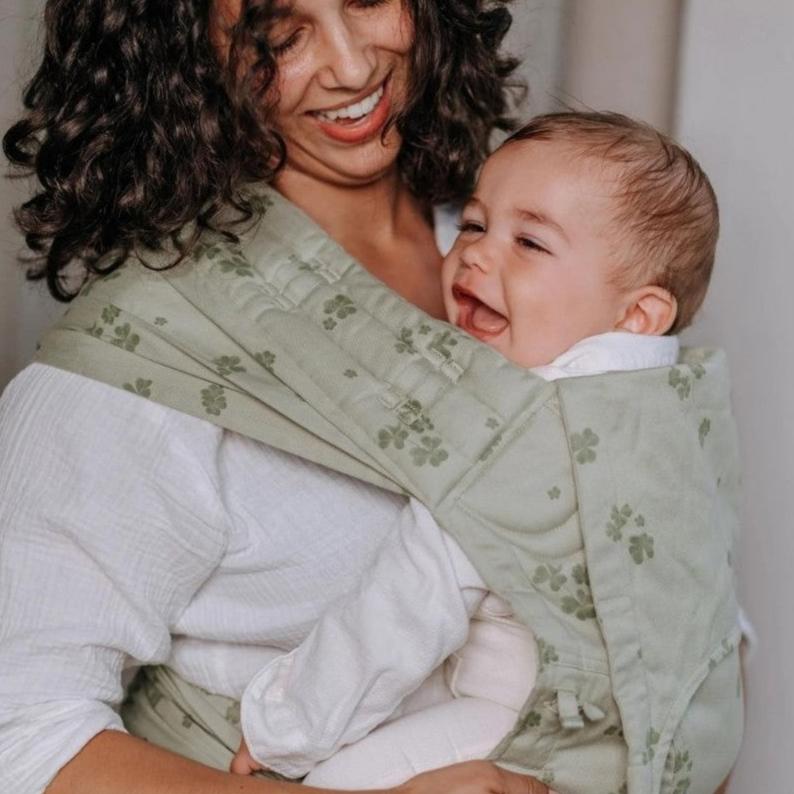 Mulher sorridente segurando um bebé num porta-bebés verde com padrão.