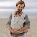 Homem com barba segurando um bebé num porta-bebés Fidella FlyTai Baby na praia.