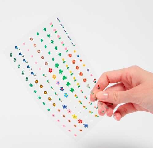 Mão a segurar uma folha de autocolantes OMY 'Flower' transparentes, com desenhos de pequenas flores e plantas.