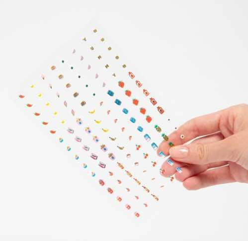 Mão a segurar uma folha de autocolantes OMY 'Yummy' transparentes, mostrando os pequenos desenhos de comida.