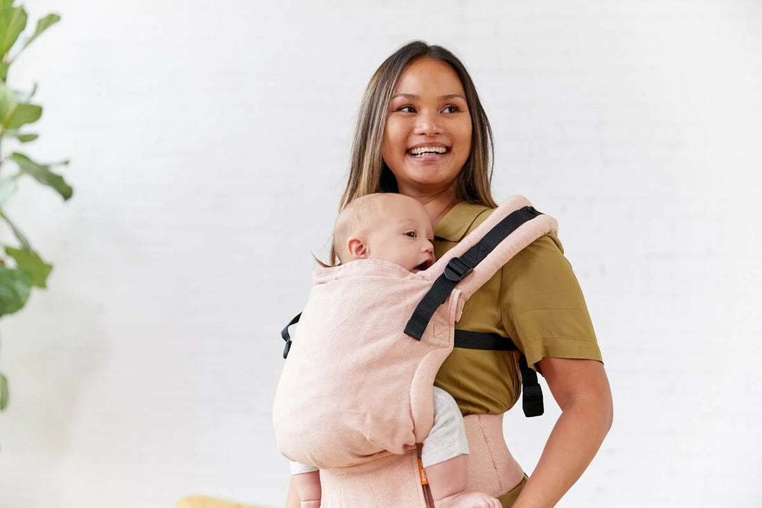 Mulher sorridente a usar a Mochila Tula Free-to-Grow Linho - Sunset, carregando um bebé pequeno na posição frontal, em ambiente interno com plantas ao fundo.