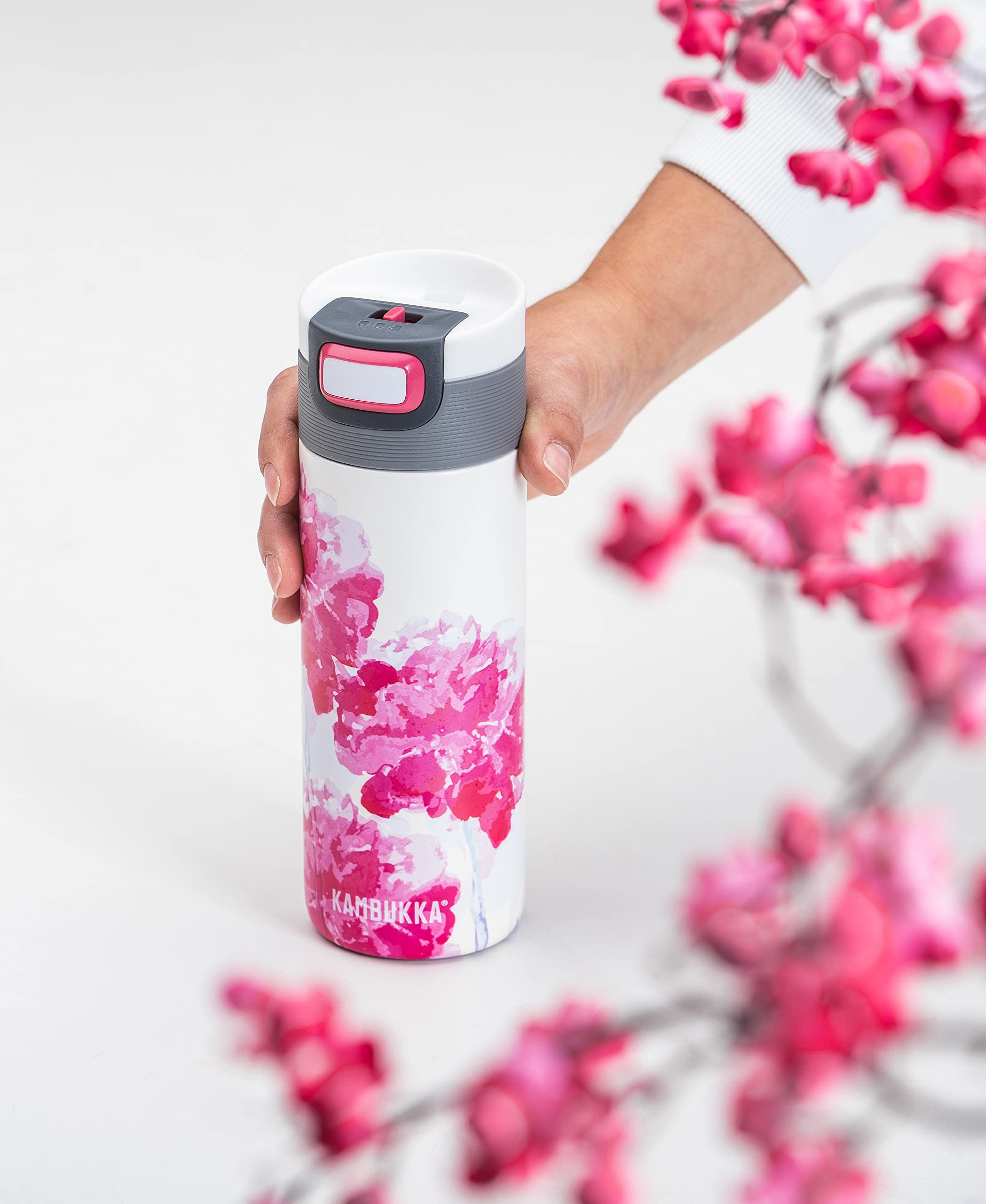 Mão a segurar a Garrafa Térmica Kambukka Etna com padrão floral rosa, destacando o design e a facilidade de uso em ambiente primaveril.