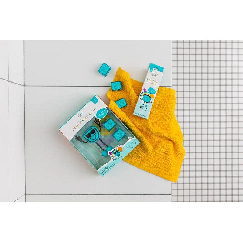 Conjunto GloPals de boneco com 2 cubos de luz (Alex Amarelo) em embalagem, junto a uma toalha amarela sobre azulejos brancos.