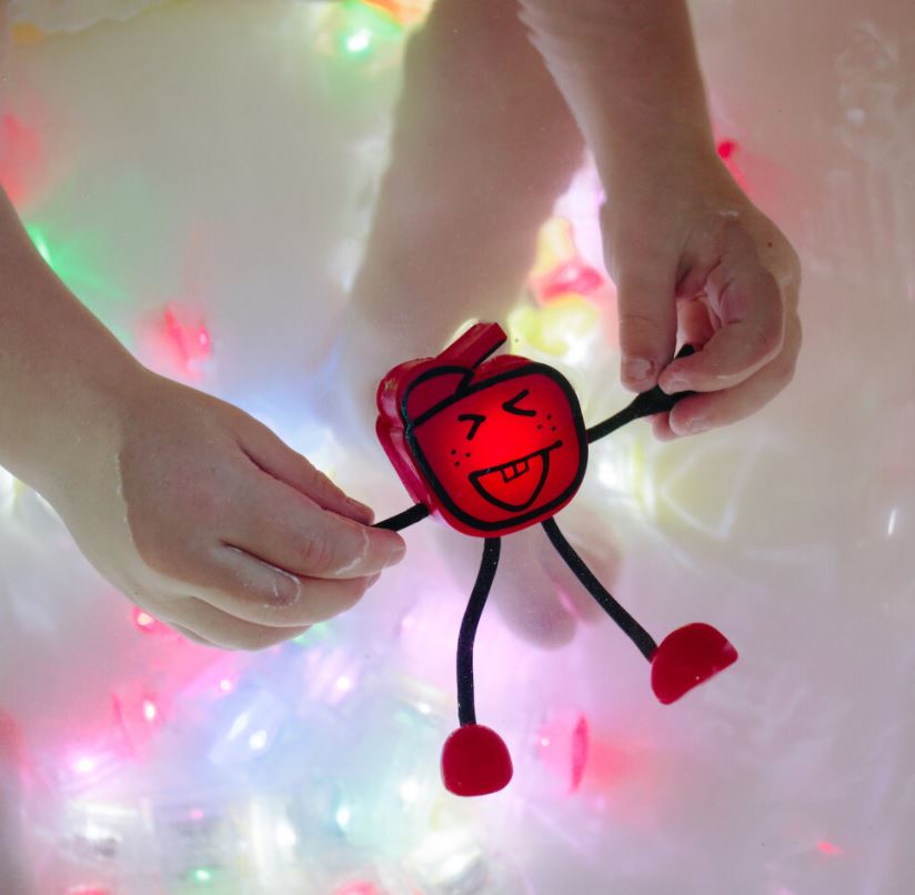 Boneco GloPals Alex amarelo a flutuar na água, iluminado por dentro com dois cubos de luz, criando um efeito mágico no banho.