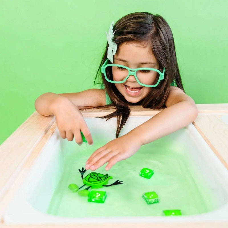 Criança sorridente com óculos verdes brinca com o boneco Glo Pal Alex (verde) e cubos de luz na água verde.
