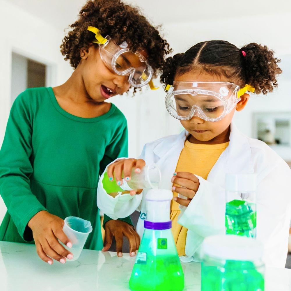 Duas crianças com óculos de proteção e batas a brincar com líquidos verdes em material de laboratório, simulando uma experiência científica.