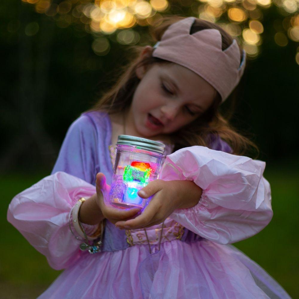 Menina vestida de princesa, com coroa, a segurar um frasco de vidro com os cubos de luz GloPals multicoloridos a brilhar no escuro.