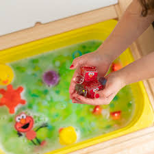 Mãos segurando cubos de luz GloPals Elmo acesos em água verde, com outros brinquedos sensoriais para banho.
