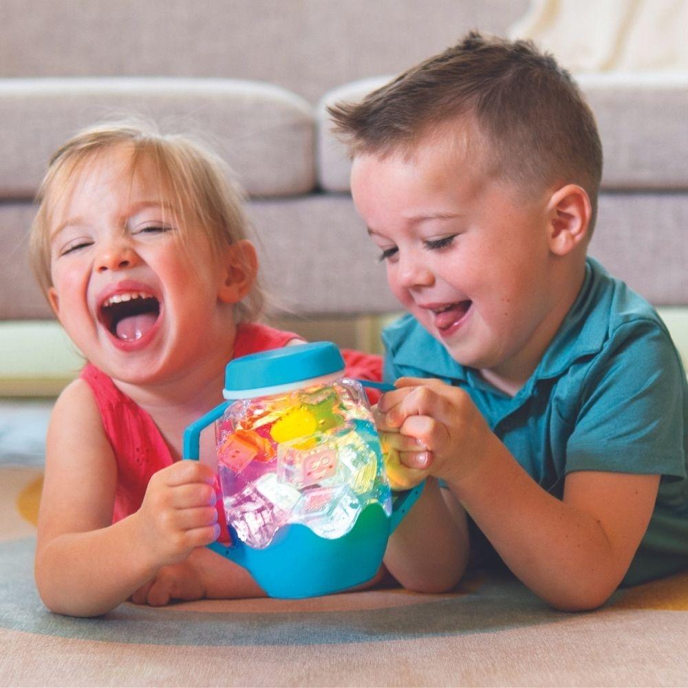 Duas crianças felizes a brincar no chão com o Jarro da Calma Sensorial GloPals azul iluminado, cheio de GloPals coloridos.