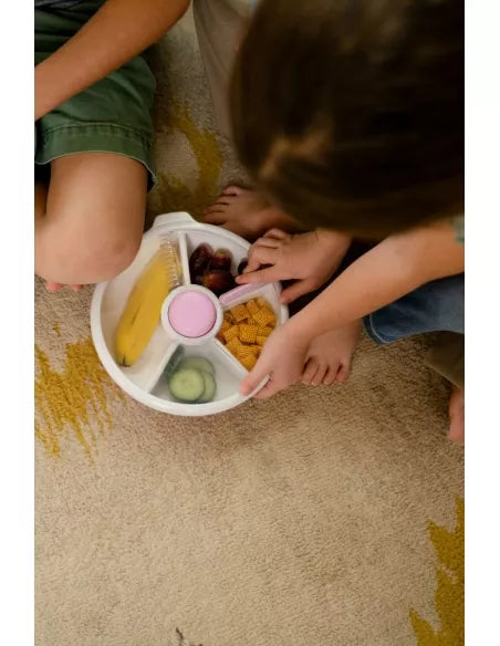 Crianças com os pés descalços a partilhar uma lancheira GoBe Snack Spin de 21 cm com banana, uvas e bolachas, num tapete.