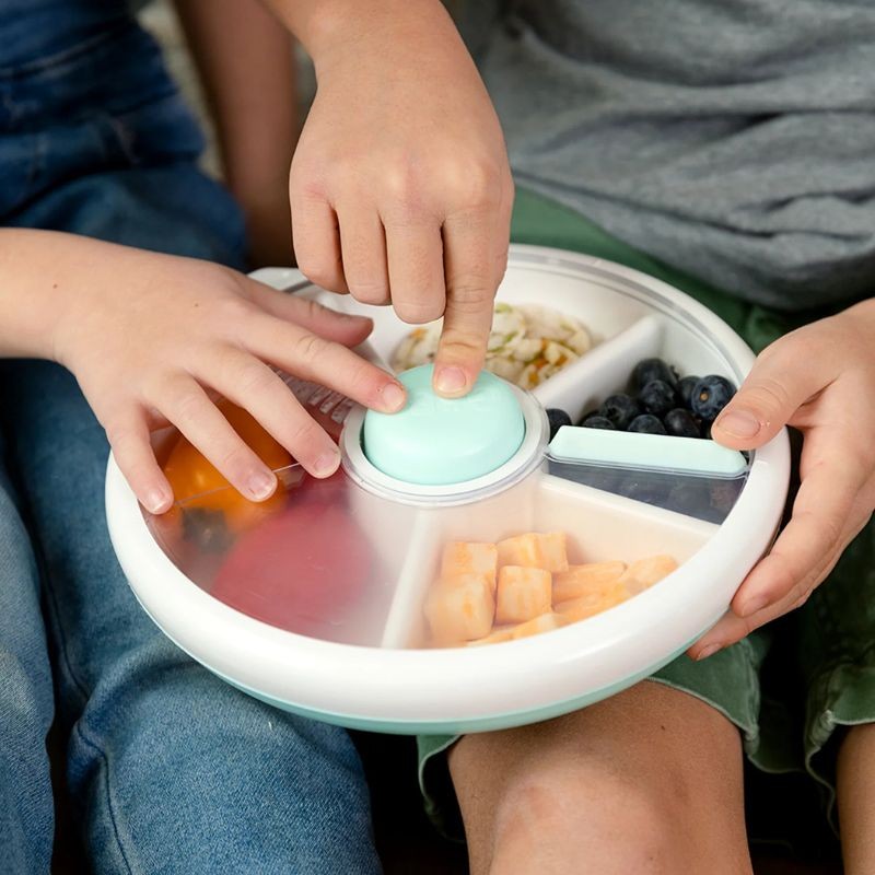 Mãos de criança a carregar na lancheira GoBe Snack Spin de 21 cm, mostrando os compartimentos com mirtilos, melão e outros lanches.