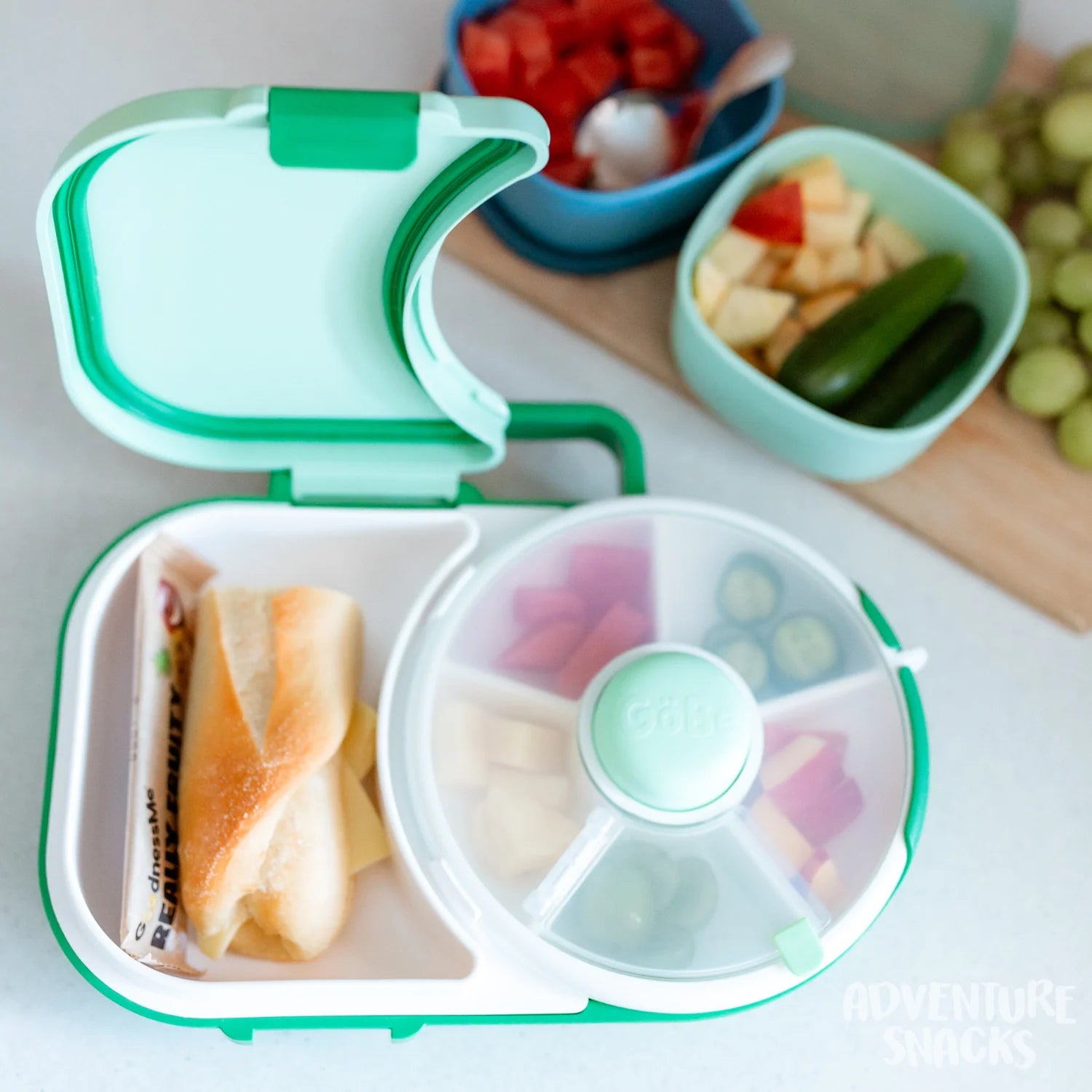 Lancheira GoBe Spin 2 em 1 aberta, verde menta, com sandes e dispensador giratório cheio de frutas e vegetais frescos.