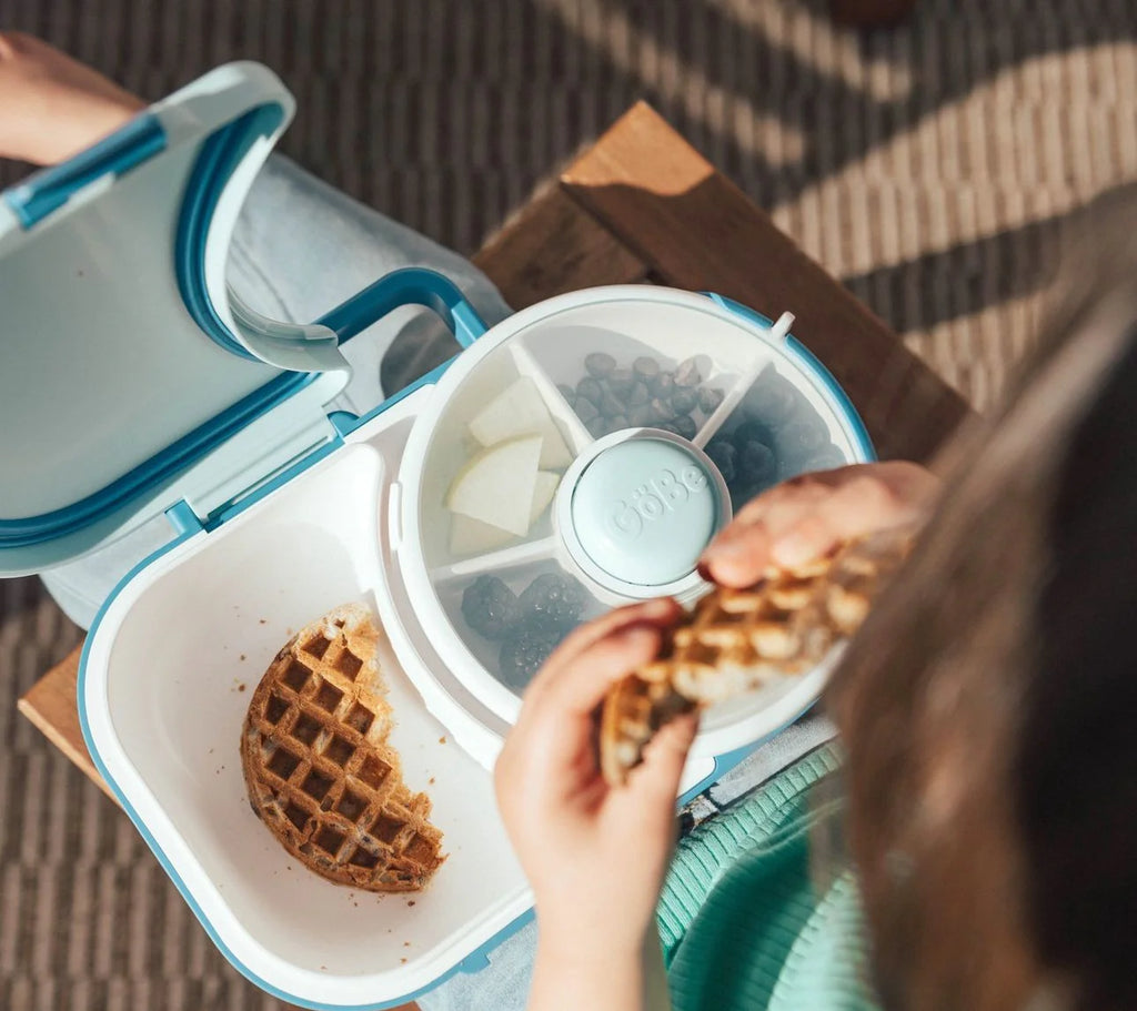 Vista superior de criança a comer um waffle do compartimento principal da lancheira GoBe, enquanto o spinner contém frutas e mirtilos.