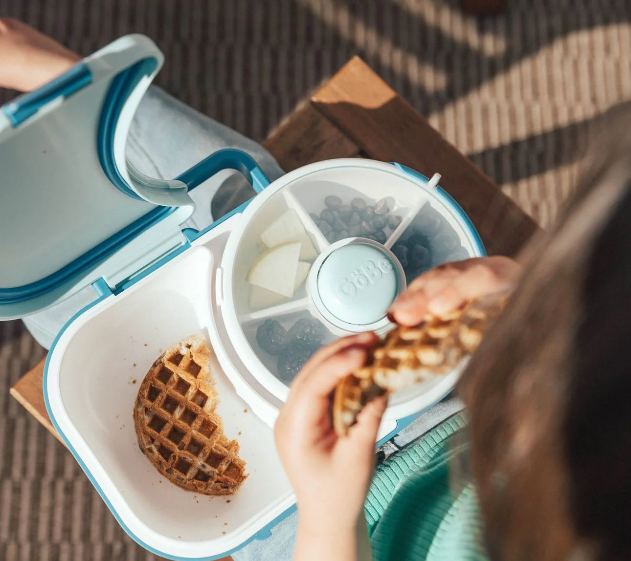 Vista superior de criança a comer um waffle do compartimento principal da lancheira GoBe, enquanto o spinner contém frutas e mirtilos.