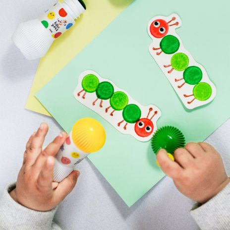 Mãos de criança usando Haku Yoka 5 dot markers para pintar lagarta com pontos verdes. Pega ergonómica fácil.