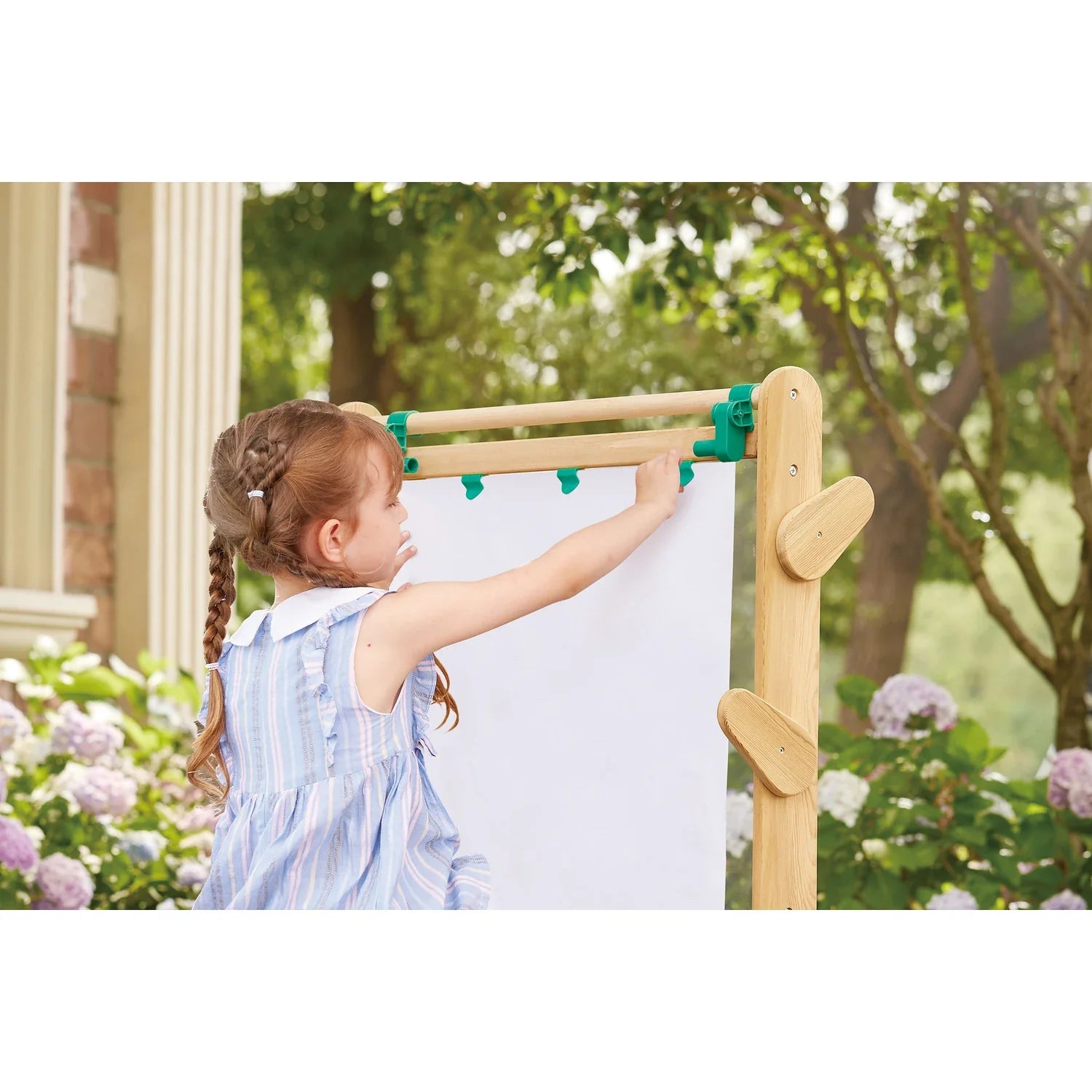 Criança a prender uma folha de papel branco no cavalete Hape de exterior, utilizando os prendedores verdes, num cenário de jardim.
