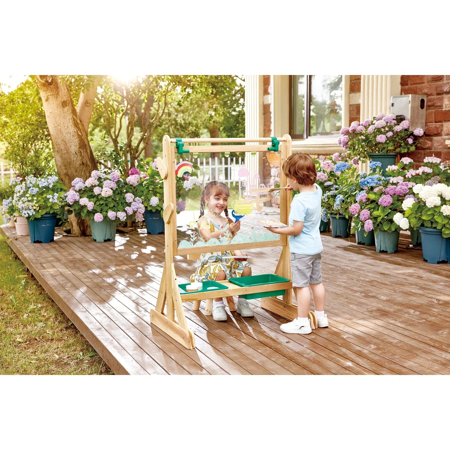 Duas crianças a brincar e a pintar simultaneamente no cavalete duplo Hape, num deck de madeira ensolarado com flores em vasos.