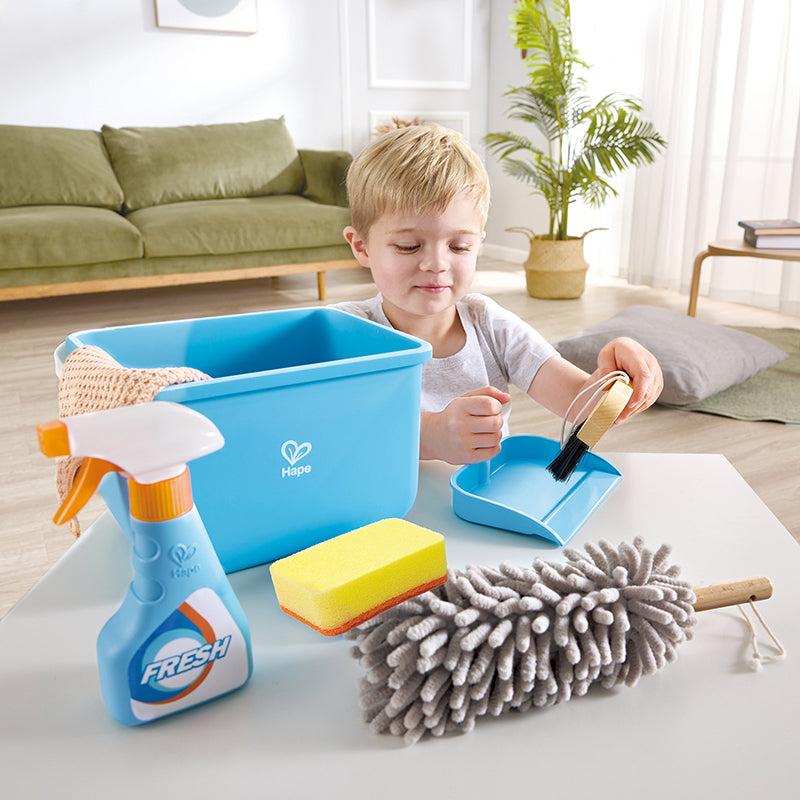 Menino brincando com o conjunto de limpeza Hape, utilizando a escova e a pá para simular a limpeza de uma mesa.