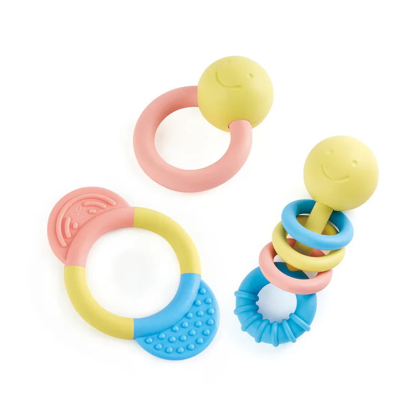 Conjunto de 3 brinquedos sensoriais Hape (guizos/mordedores) em cores pastel (rosa, amarelo, azul) sobre fundo branco.