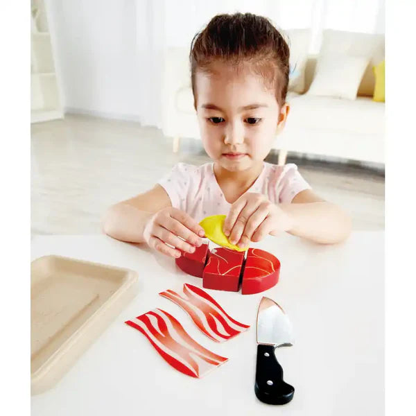 Criança a brincar com o conjunto de comida de madeira Hape, colocando uma fatia de limão sobre o bife cortado, com faca e bacon visíveis.