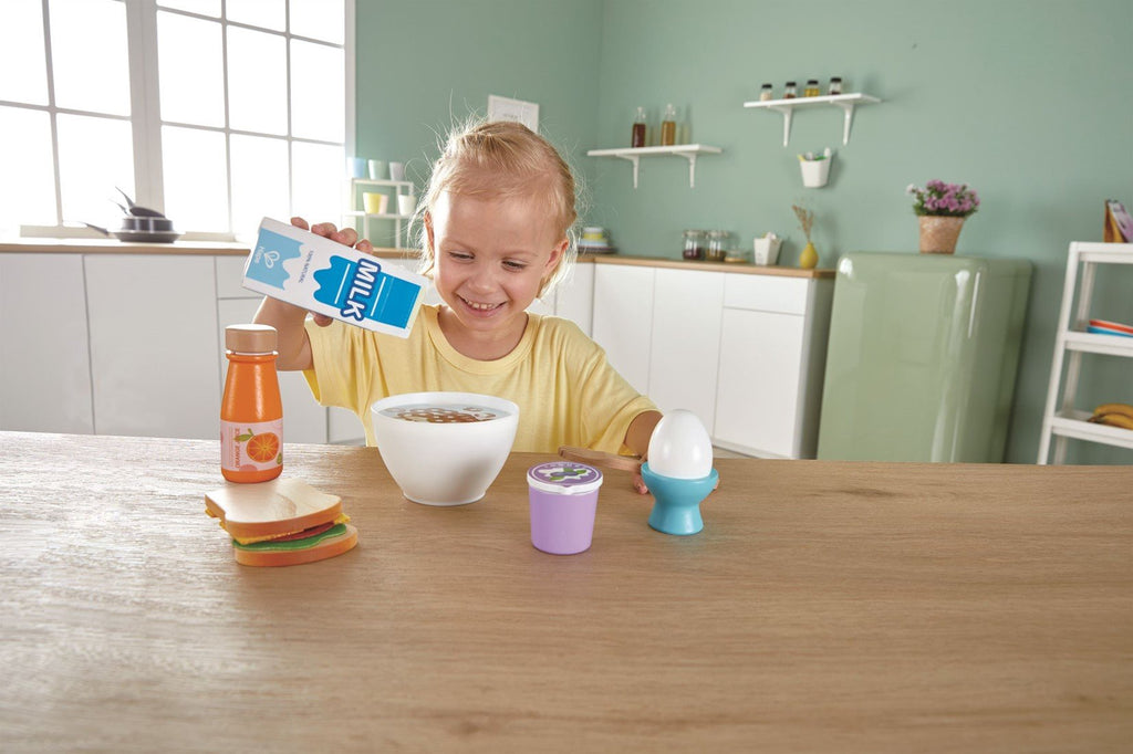 Criança feliz a brincar com o conjunto de pequeno-almoço Hape, servindo leite numa taça, com sanduíche e iogurte na mesa.