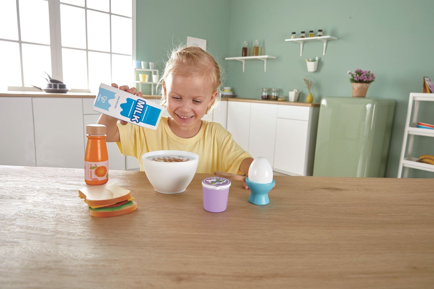 Criança feliz a brincar com o conjunto de pequeno-almoço Hape, servindo leite numa taça, com sanduíche e iogurte na mesa.
