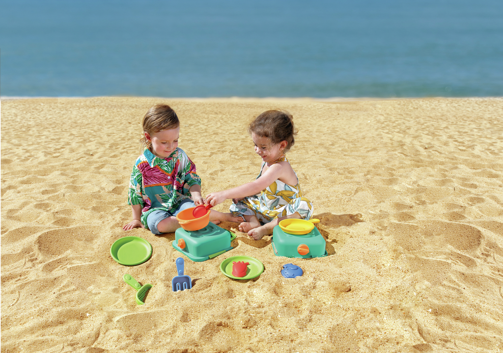 Duas crianças a brincar com o conjunto de cozinha Hape na areia da praia.