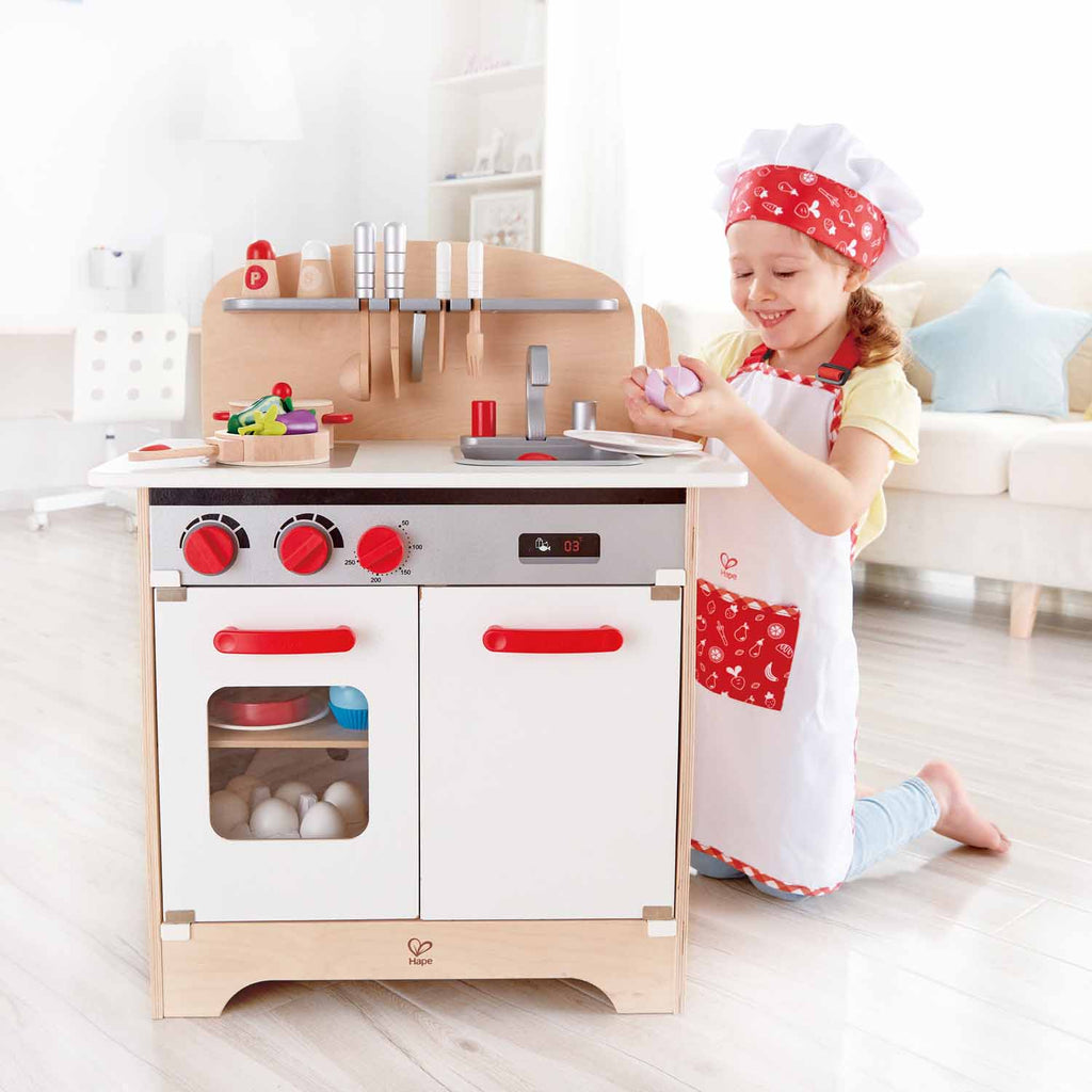 Menina com o Hape Fato de Chef pastelaria a brincar numa cozinha de faz de conta.