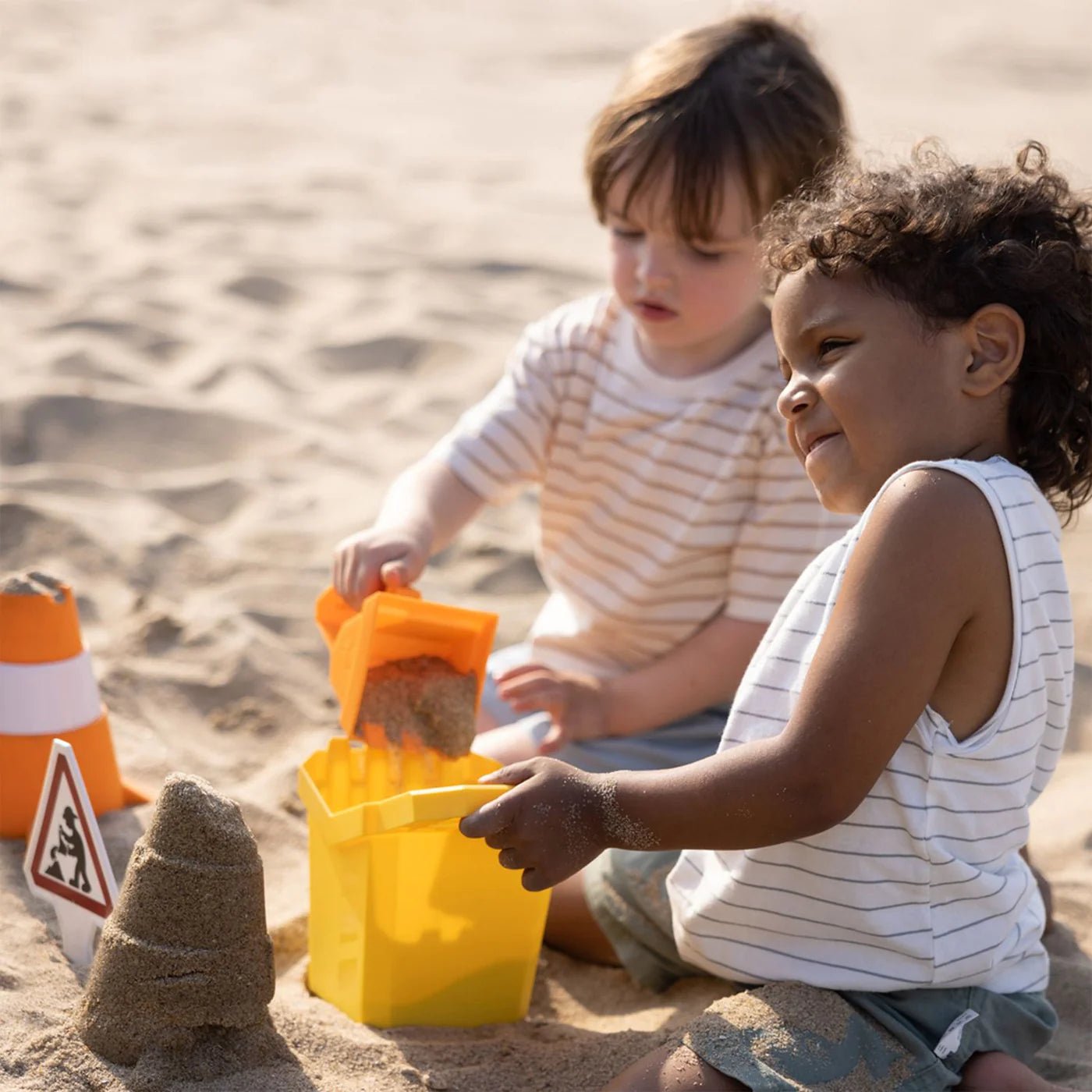 Duas crianças a brincar na areia com o set de brinquedos de praia Hape, construindo um castelo e usando o balde e pá.