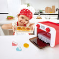 Criança sorridente com chapéu de chef apresenta uma travessa com biscoitos de madeira ao lado do Forno Mágico Hape vermelho e branco.