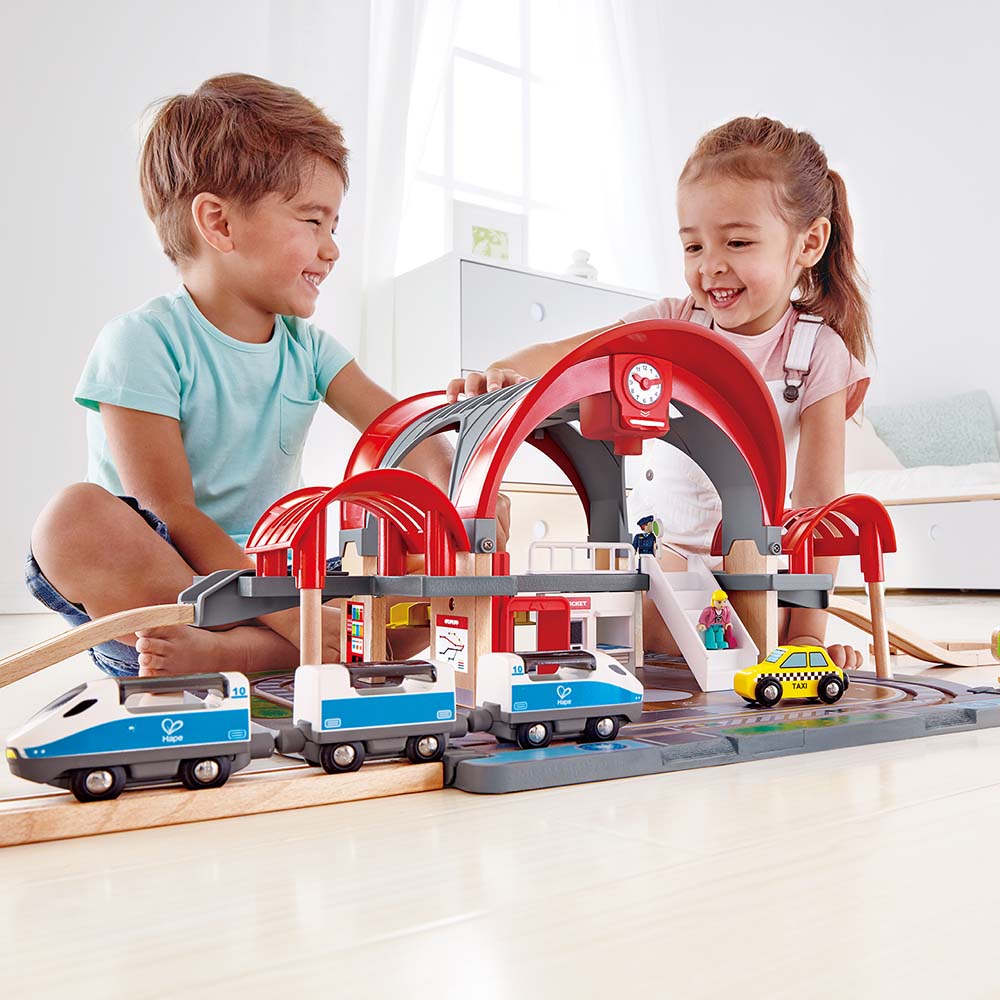 Duas crianças a brincar alegremente com o Hape Mega Kit Construção, comboios e estação montados no chão.