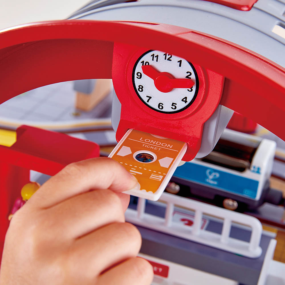 Detalhe do Hape Mega Kit: mão a inserir bilhete de comboio "London Ticket" no dispensador com relógio da estação.