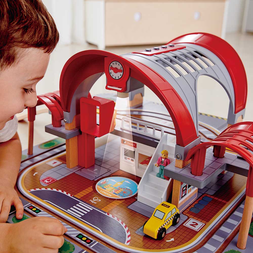 Criança a observar o Hape Mega Kit Construção, focando na estação com relógio, luz e figuras de passageiros.