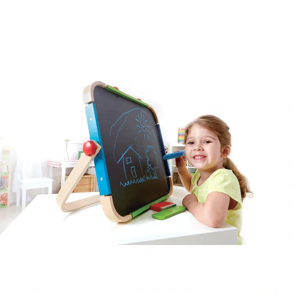 Criança sorridente a desenhar uma casa no quadro negro do cavalete Hape portátil com giz azul.