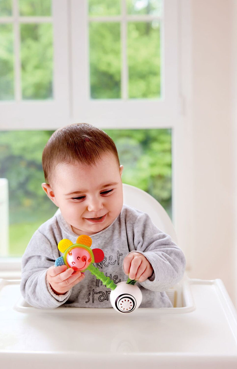 Bebé sentado numa cadeira de papa, a observar e manipular o Hape Guizo com luz e formas com expressão de curiosidade.