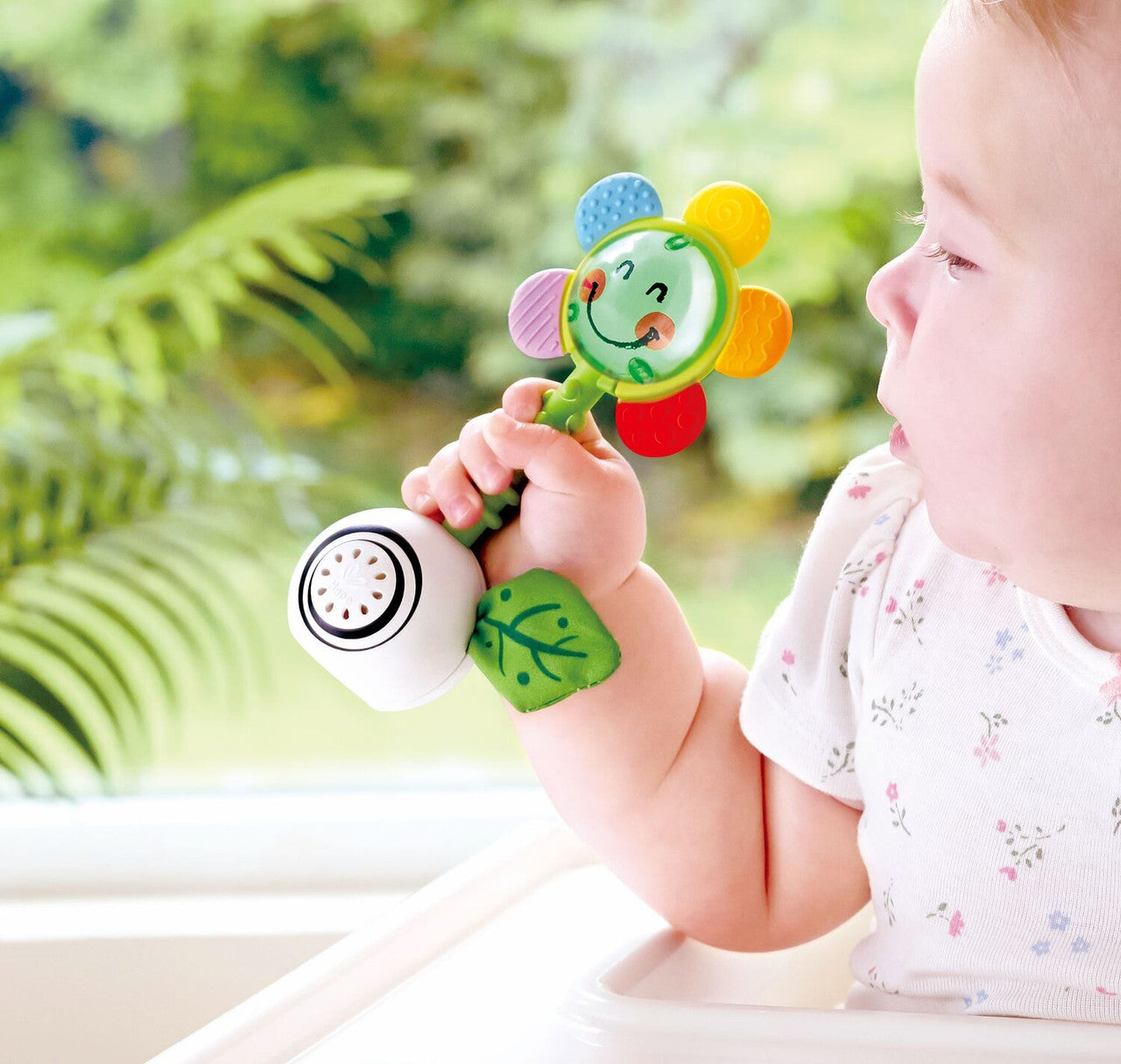 Bebé a segurar o Hape Guizo com luz e formas, com foco nas texturas das pétalas e o design ergonómico.