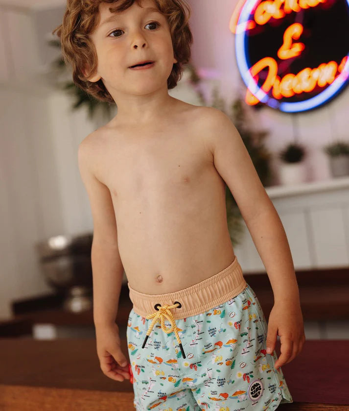 Criança de cabelo encaracolado a usar calções de banho Hello Hossy "New Zealand" com padrão temático e cós elástico bege com cordão amarelo.