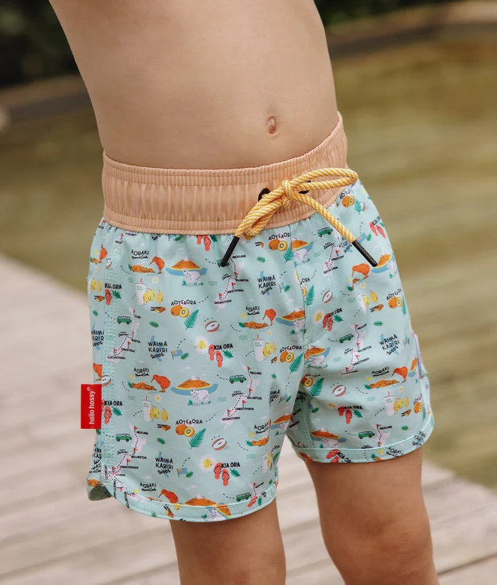 Calções de banho infantis Hello Hossy "New Zealand" com padrão ilustrado, vestidas por uma criança, destacando o cós elástico bege e o cordão amarelo.