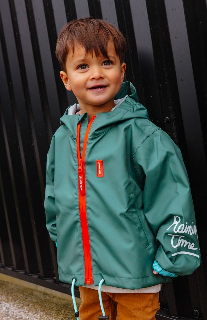 Detalhe do Casaco Captain impermeável verde da Hello Hossy, mostrando o capuz, fecho laranja e inscrição "Raining Time" na manga.