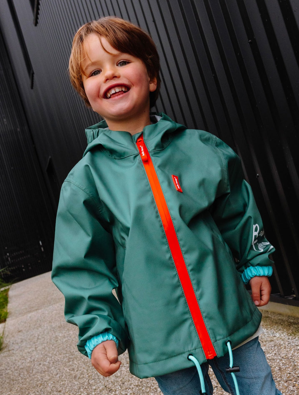 Criança sorridente vestindo o Casaco Captain impermeável verde-azulado da Hello Hossy com fecho laranja vibrante.