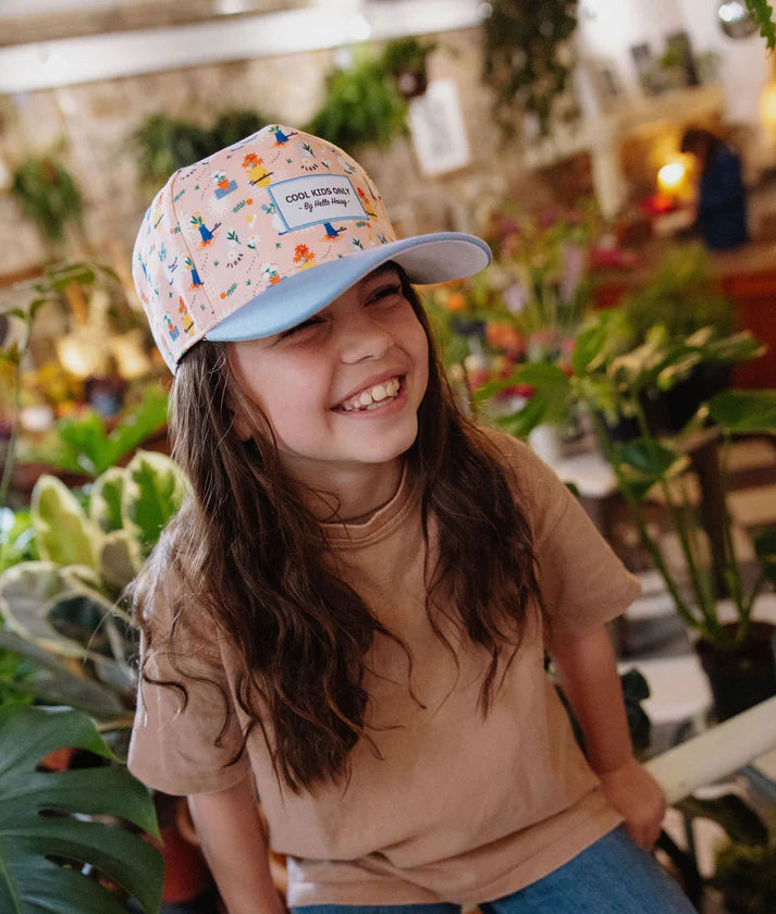 Criança sorrindo usando o boné Flore Hello Hossy. Padrão floral e aba plana em destaque. Acessório moderno e divertido.