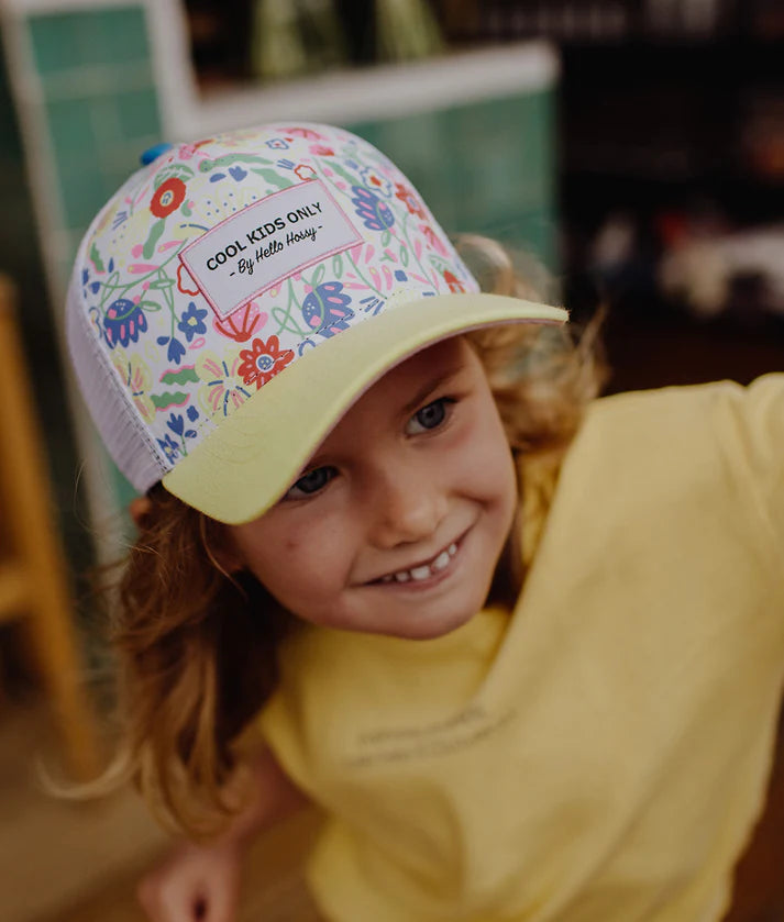 Criança sorridente usa boné infantil Hello Hossy Garden Party com padrão floral colorido e aba amarela.