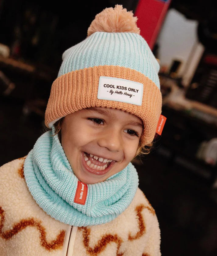 Criança sorridente a usar o gorro HELLO HOSSY Candy azul-claro e salmão com pompom e cachecol a condizer.