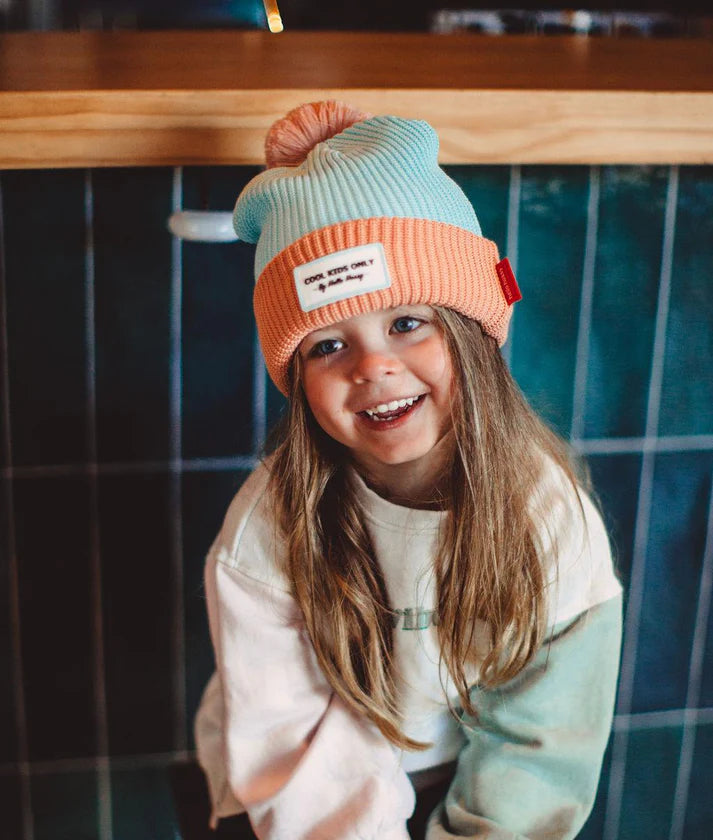 Menina com cabelo comprido a usar o gorro HELLO HOSSY Candy azul e salmão, sorrindo em frente a um fundo de azulejos verdes.