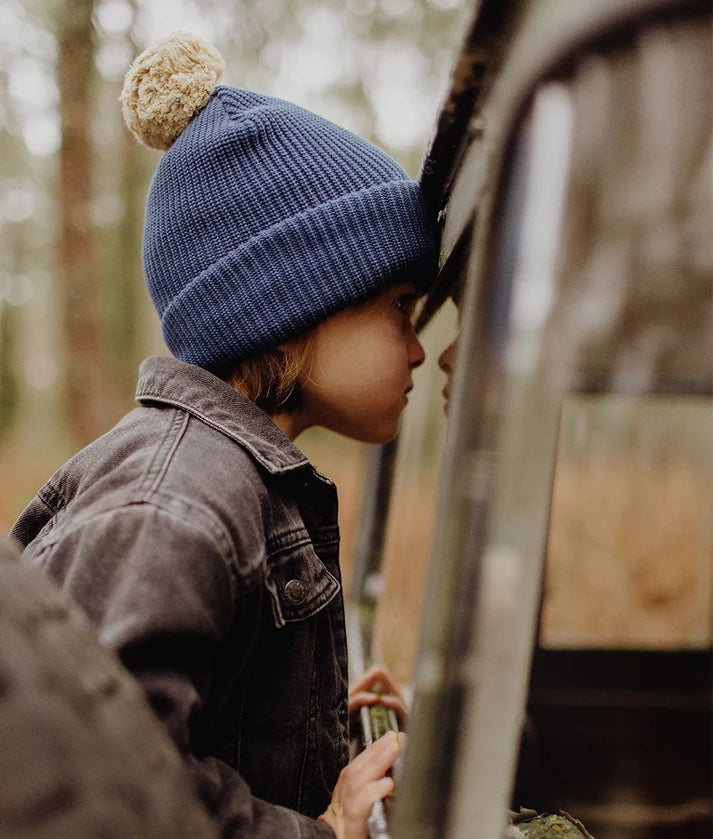 Criança com gorro azul-escuro Hello Hossy e pompom bege a olhar pela janela de um veículo no exterior.