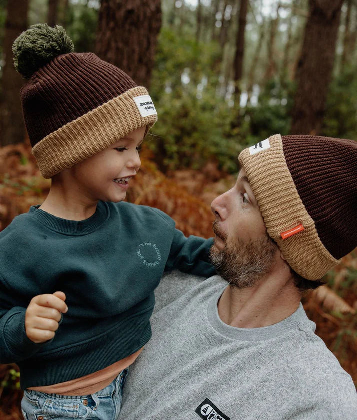 Close-up do pai e filho a olharem-se, ambos com o gorro de malha canelada castanho e bege da Hello Hossy, com pompom.