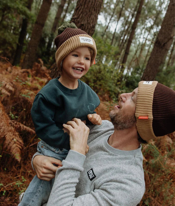 Criança sorridente ao colo do pai, ambos a usar gorros Hello Hossy castanho e bege com pompom verde, num cenário de floresta.