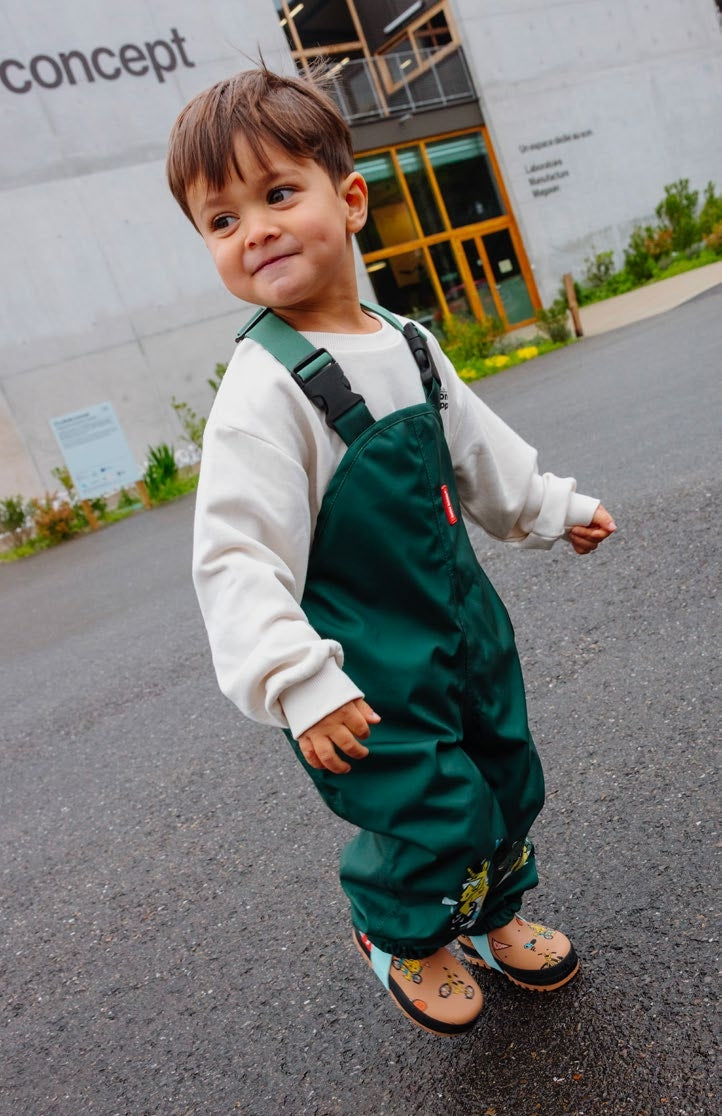 Criança sorridente a usar o Macacão Captain verde escuro da Hello Hossy, ideal para brincadeiras ao ar livre.
